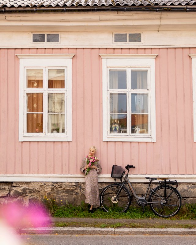 Nainen polkupyörän kanssa vaaleanpunaisen vanhan puutalon edessä.